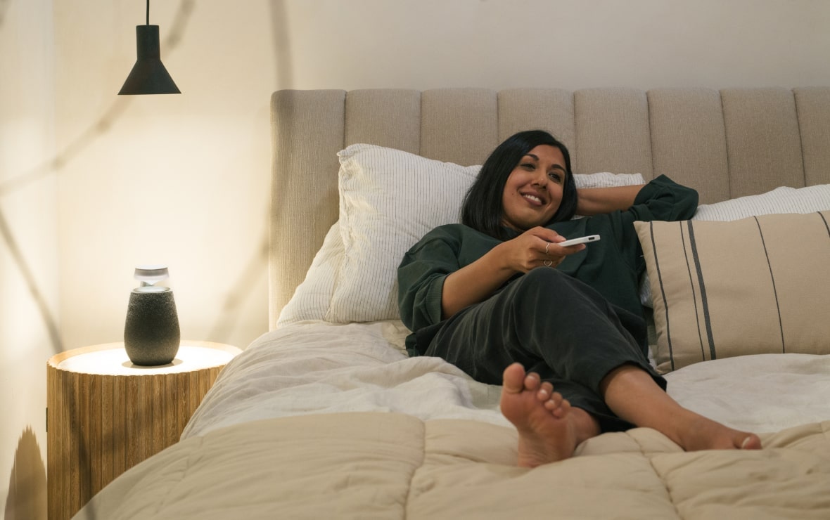 Lisa Mistry liegt im Bett und benutzt die Fernbedienung neben dem LG Xboom XO2T mit aktivem Mood Lighting.