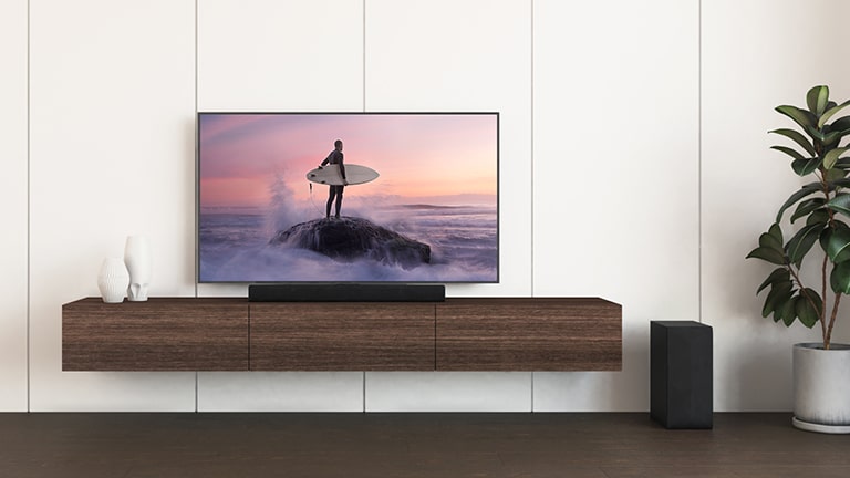 Et LG TV og en LG Soundbar er placeret på en brun hylde, og subwooferen er på gulvet. Tv-skærmen viser en surfer, der står på klippen.