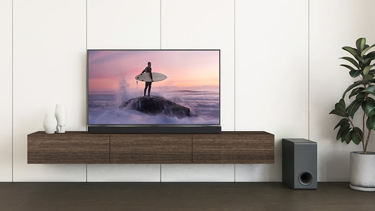 Et LG TV og en LG Soundbar er placeret på en brun hylde, og subwooferen er på gulvet. Tv-skærmen viser en surfer, der står på klippen.