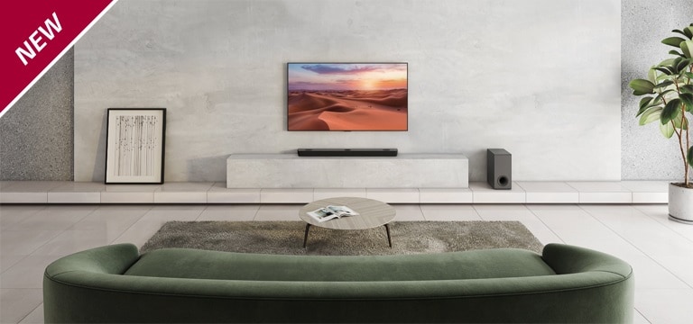 TV'et hænger på den hvide søjle i midten af stuen. På den hvide hylde nedenunder er der placeret en LG Sound Bar. På gulvet lige under soundbaren står der en sort subwoofer. Bag den grønne sofa er der placeret to baghøjttalere på sofabordet i både venstre og højre side. NYT-mærke vises i øverste venstre hjørne.