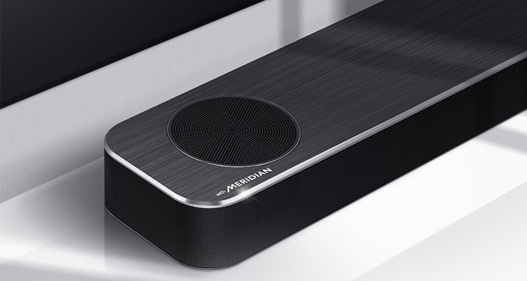 Nærbillede af venstre side af en LG Soundbar med et Meridian-logo vist nederst i venstre hjørne på et produkt. Nederste venstre side af tv’et er også synligt.