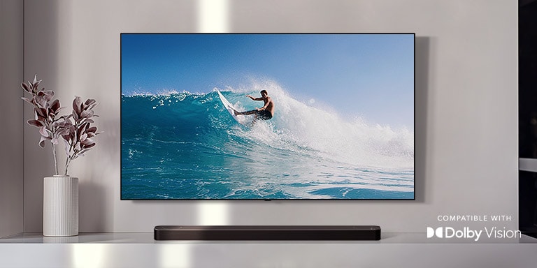 Tv på væg. Et tv viser en mand, der surfer på en stor bølge. LG Soundbar er lige under et tv på en hvid hylde. Der er en vase med en blomst lige ved siden af soundbaren. (afspil videoen)