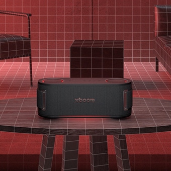 LG XBOOM-højttaler placeret på et bord i et rum med rødtoner, vægge med gittermønster og moderne møbler.