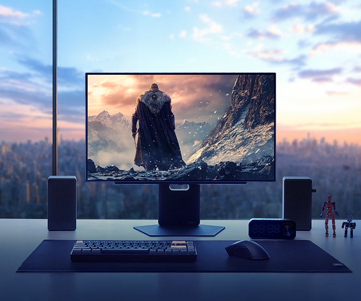En professionel gaming-monitor viser en dramatisk fantasy-spilscene med en kappeklædt kriger, som står foran en sneklædt bjergkæde. Skærmen står på et elegant skrivebord i en højhuslejlighed med en skyline ved solnedgang i baggrunden. Skærmen er omgivet af højttalere, et tastatur, et headset, et ur og små actionfigurer, hvilket fremviser en fordybende og stilfuld gaming-opsætning.