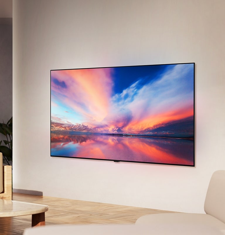 LG OLED TV، OLED B4 على جدار غرفة معيشة محايدة الألوان يعرض صورة ملونة لمنظر غروب الشمس فوق المحيط.