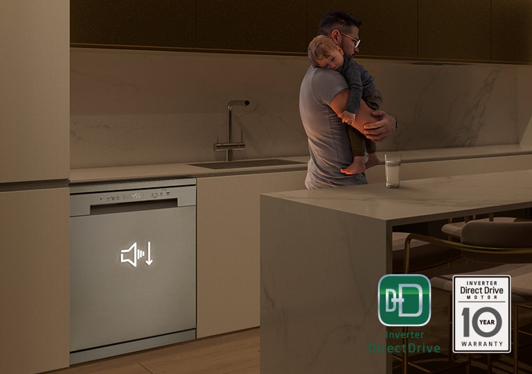 Man holding sleeping baby in a dimly lit kitchen, free-standing dishwasher operating quietly in the background.