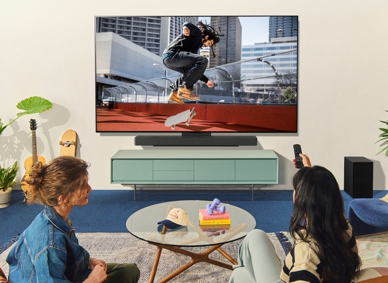 Two women sitting on a sofa in front of a wall-mounted LG TV in a colorful living room, and one woman points a remote at the TV screen. 