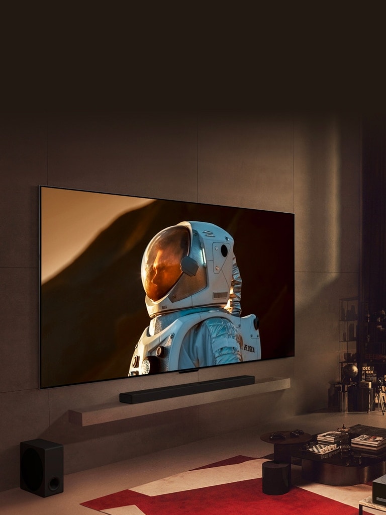 An ultra-large LG TV mounted on a wall in a cozy living room displaying a close-up shot of astronaut takes up the majority of the wall. A man sits on a sofa in front watching the TV with a dog, and a night cityscape is beyond the window.