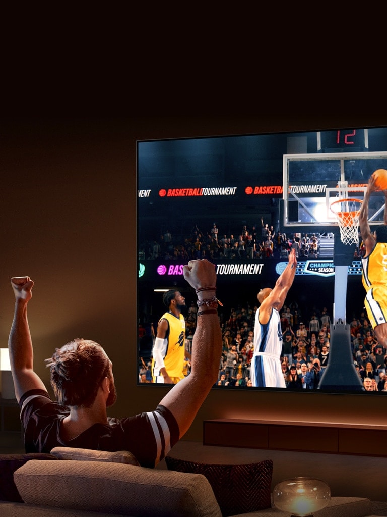 A man sits on a sofa in front of an ultra-large LG TV displaying a basketball game, with his arms in the air as if cheering.