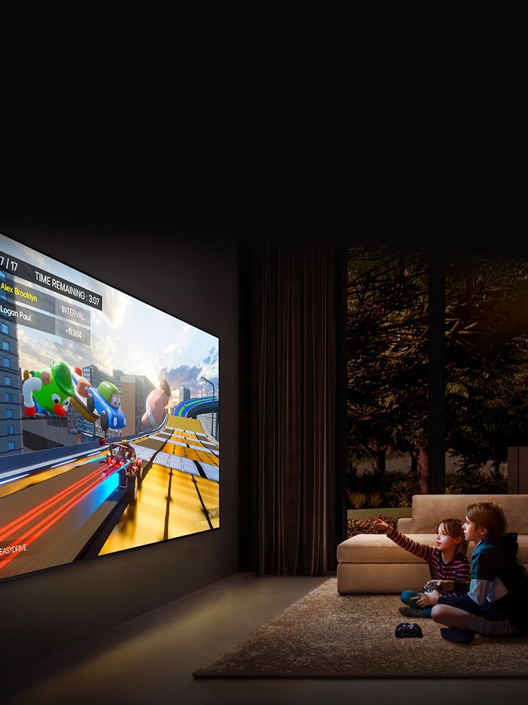 A side-angle view of an ultra-large LG TV mounted on a wall in a cozy living room displaying a colorful game, and two children sitting on the floor in front of the TV holding game controllers.