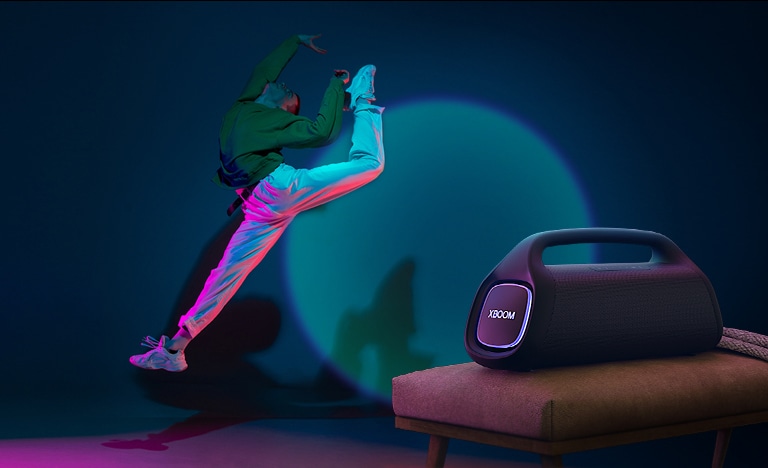LG XBOOM Go XG9 está colocado sobre la silla proyectando su iluminación de escenairo en la pared y una persona está bailando frente a ella.