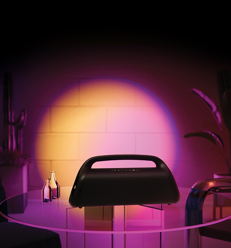 Una imagen de LG XBOOM Go XG9 colocado en una mesa de cristal en casa, con una iluminación de escenario naranja y rosa. 