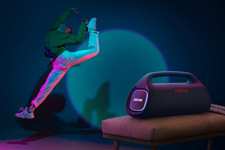LG XBOOM Go XG9 está colocado sobre la silla proyectando su iluminación de escenairo en la pared y una persona está bailando frente a ella.
