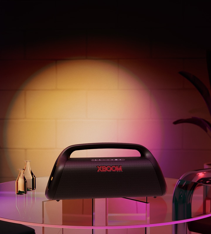 Una imagen de LG XBOOM Go XG9 colocado en una mesa de cristal en casa, con una iluminación de escenario naranja y rosa. 