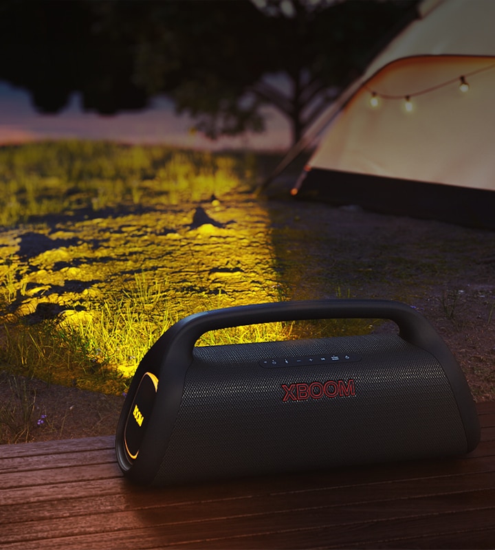 LG XBOOM Go está colocado sobre un suelo de madera proyectando una luz de escenario en un camping.