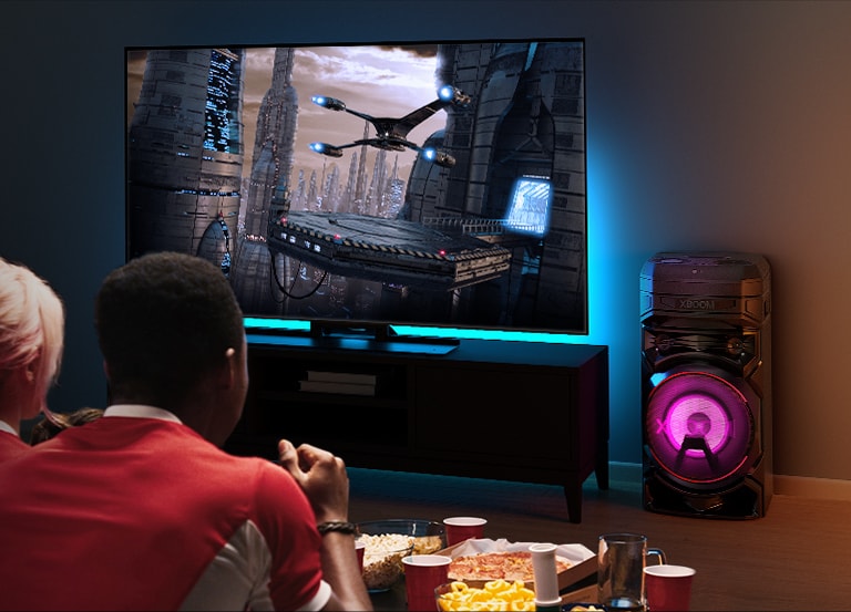 Gente viendo la TV en una pared con una LG XBOOM RNC5 a su derecha. El televisor muestra una película.