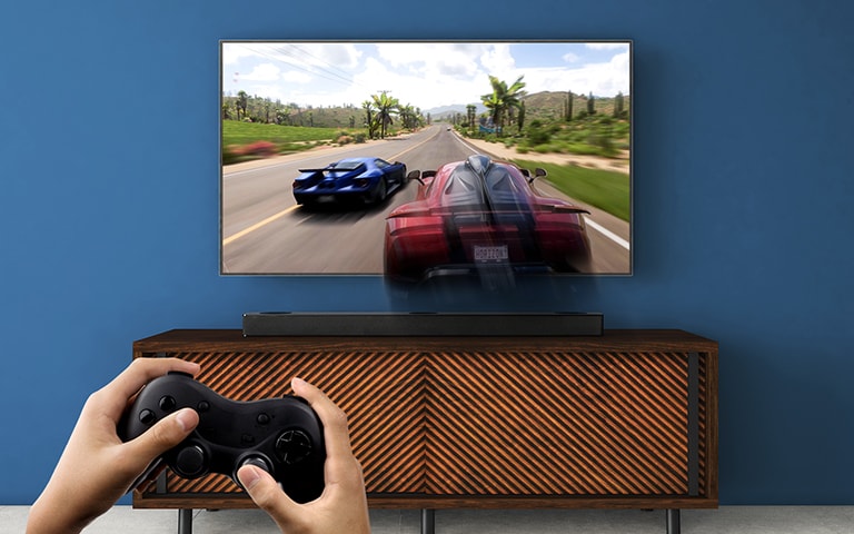 LG TV está en la pared, mostrando un juego de carreras. La barra de sonido LG se coloca en el estante marrón, justo debajo del televisor LG. Un hombre sostiene un joystick.