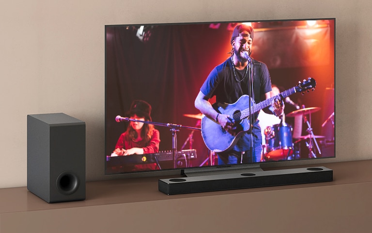 El televisor LG muestra un concierto y la barra de sonido LG se coloca debajo del televisor LG. A la izquierda, el subwoofer está en el estante marrón.