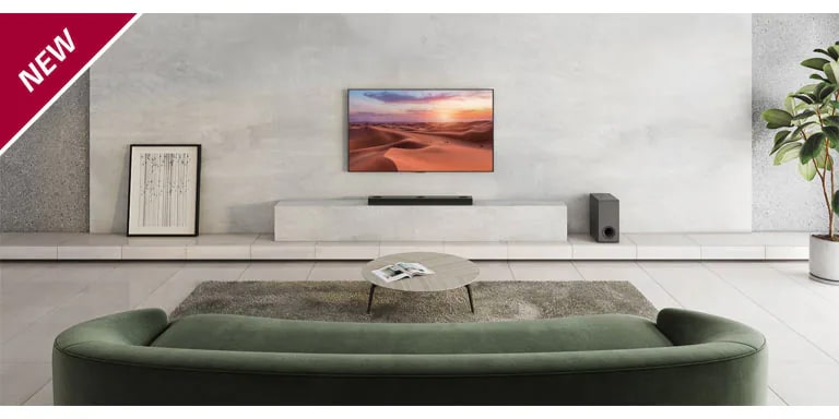 El TV está colgado en la columna blanca del centro del salón. Debajo, la barra de sonido LG está colocada en la estantería blanca. En el suelo, justo debajo de la barra de sonido, hay un subwoofer inalámbrico. Detrás del sofá verde, se han colocado dos altavoces traseros encima de unas mesitas de centro, una en cada lado. La marca NEW se visualiza en la esquina superior izquierda.