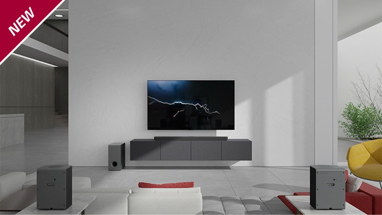 La barra de sonido está colocada en un mueble gris con un TV colgado en la pared de un salón. Un subwoofer inalámbrico está colocado en el suelo, en el lado izquierdo, y la luz del sol entra por el lado derecho de la imagen. Un sofá largo de color blanco y rojo está colocado frente al televisor y la barra de sonido. La marca NEW se visualiza en la esquina superior izquierda.