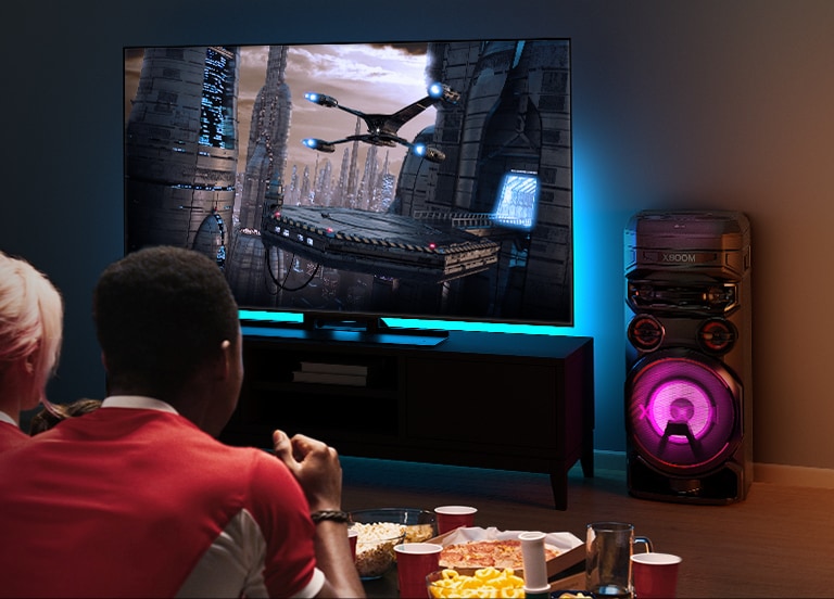 Gente viendo la TV en una pared con una LG XBOOM RNC7 a su derecha. El televisor muestra una película.
