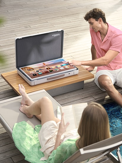 El StanbyME Go está colocado sobre la mesa del patio, y la pantalla está en modo mesa, mostrando la pantalla de inicio. Un hombre está a punto de tocar una de las aplicaciones, mientras una mujer se relaja.