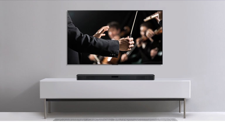 Vemos un TV en una pared gris con la Barra de Sonido LG debajo en una balda gris. En el TV un director dirige a su orquesta.