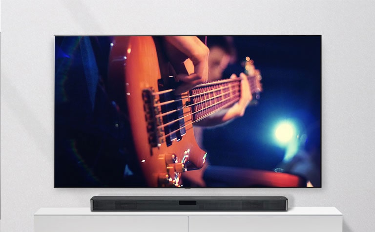 El TV está inslatado sobre pared y la barra de sonido sobre una balda blanca. El TV muestra aun hombre tocando la guitarra.