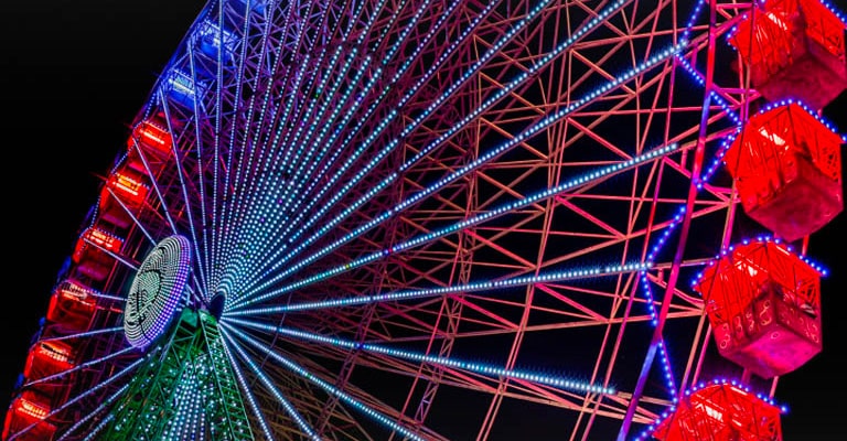 Una rueda de la fortuna ligeramente descolorida se ilumina por la noche, con destellos de colores que comienzan en el lado izquierdo de la pantalla y se mueven a través de ella. A medida que pasan los destellos de colores, la pantalla pasa de nublada a colores brillantes y vivos.