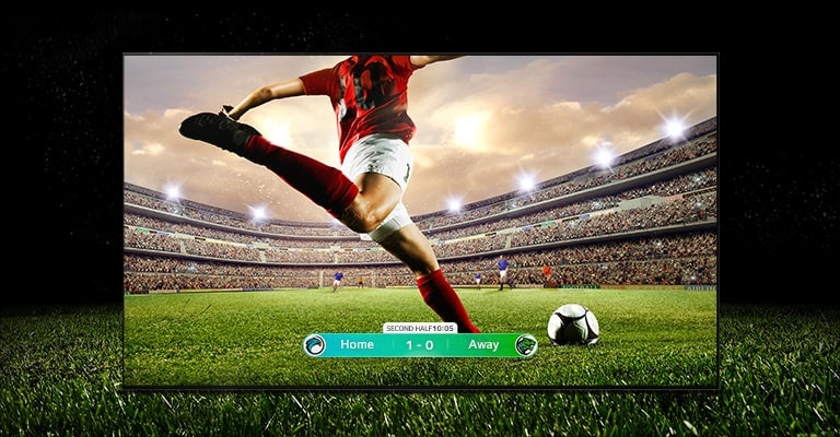 Imagen de una pantalla que muestra un partido de fútbol con un jugador vestido de rojo a punto de chutar el balón. En la parte inferior de la pantalla se ve el marcador del partido. La hierba verde del campo se extiende más allá de la pantalla hasta el fondo negro.