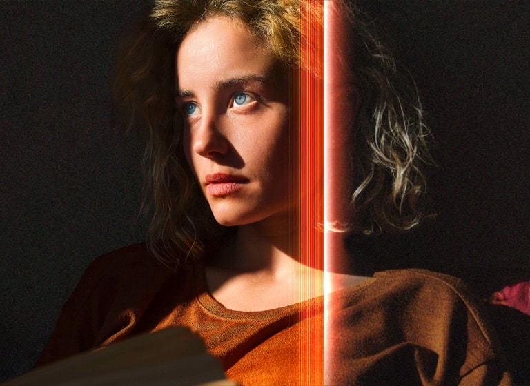 Una mujer con penetrantes ojos azules y un top naranja quemado en un espacio oscuro. Líneas rojas que representan refinamientos de AI cubren parte de su rostro, que es brillante y detallado, mientras que el resto de la imagen parece apagada.