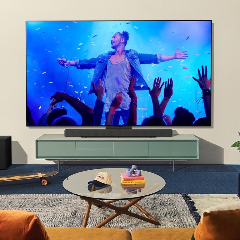 Se muestra, en un salón moderno, un TV OLED y una barra de sonido que se combinan con el soporte Synergy