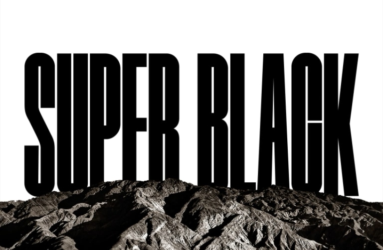 Las palabras “SUPER BLACK” aparecen en mayúsculas resaltadas negras. Una escena con montañas negras, con una definición nítida, se eleva tapando las letras, y revelando un pueblo y unas dunas de arena. La copia negra desaparece detrás de un cielo negro.