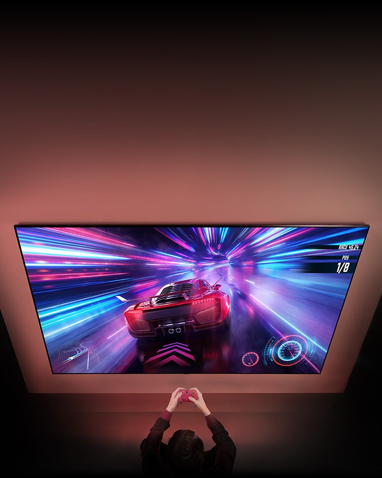 There is a big TV on the wall and you can see the racing game screen in the screen. In front of the TV, you can see the hands and controllers of the person who focuses on the game.