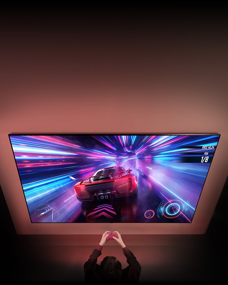 Hay un TV grande en la pared y en la pantalla se puede ver un juego de carreras de coches. Delante del TV, se pueden ver las manos y los controladores de la persona que juega.
