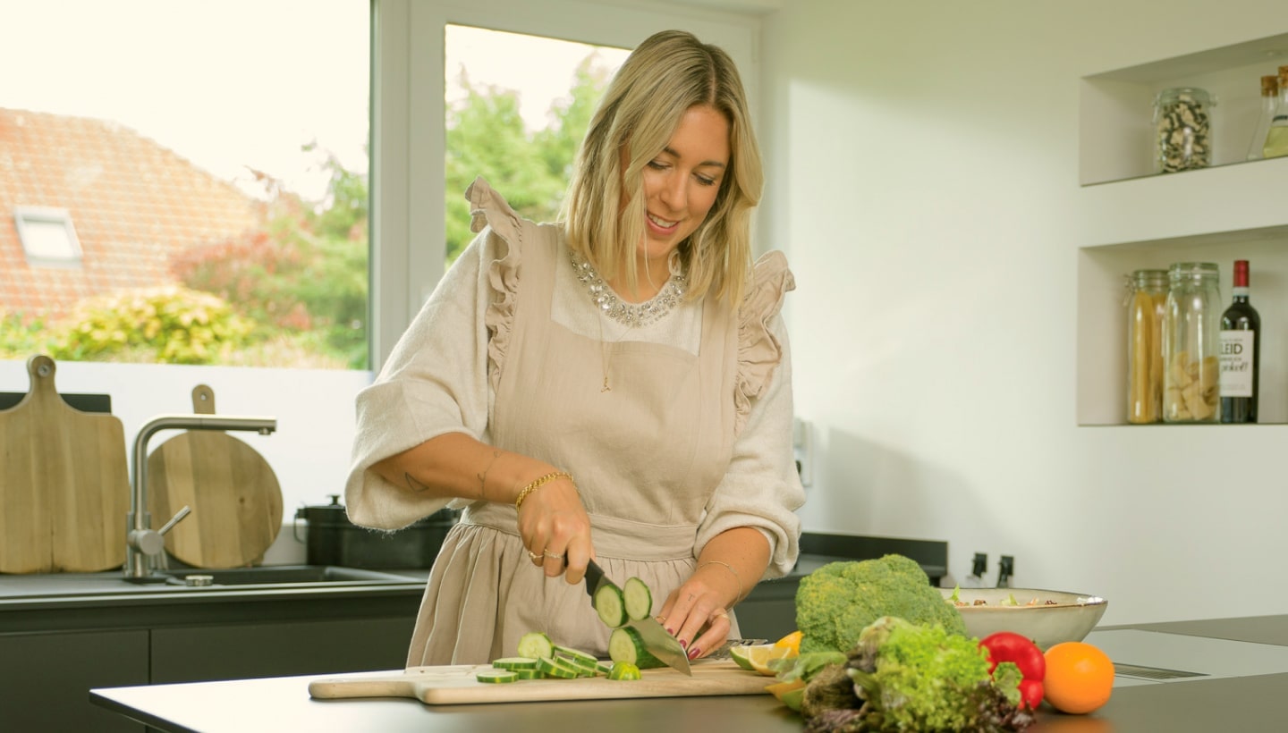 Una mujer corta pepinos frescos en una cocina luminosa con verduras coloridas en la encimera.