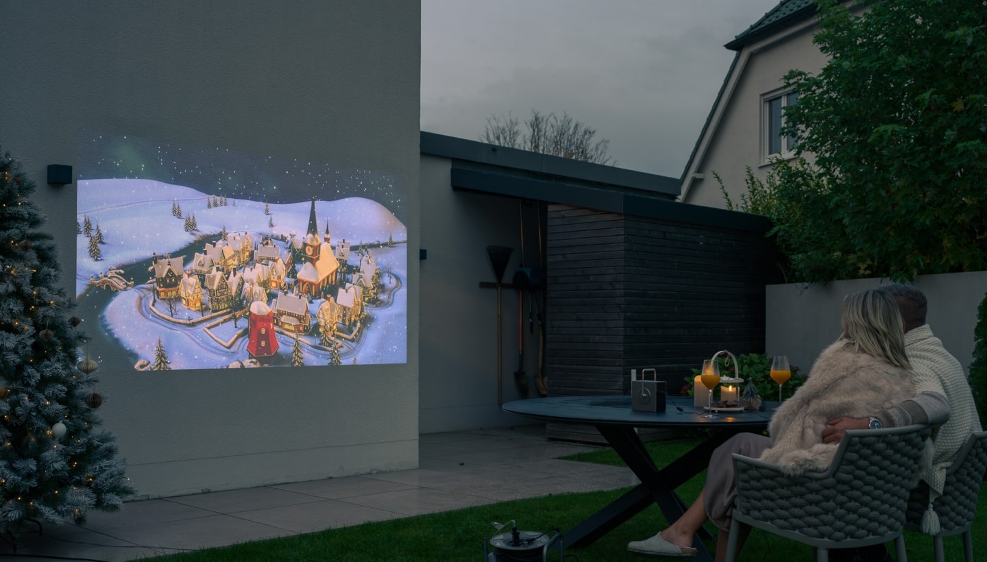 Una pareja disfruta de una noche de cine al aire libre con un proyector LG CineBeam que muestra una escena invernal festiva en una pared.