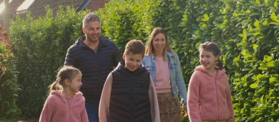 Familia de cinco personas disfrutando de un paseo al aire libre, sonriendo y vestida con ropa casual, rodeada de setos verdes.