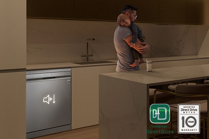 Hombre con un bebé dormido en brazos en una cocina poco iluminada, con un lavavajillas autónomo funcionando silenciosamente al fondo.