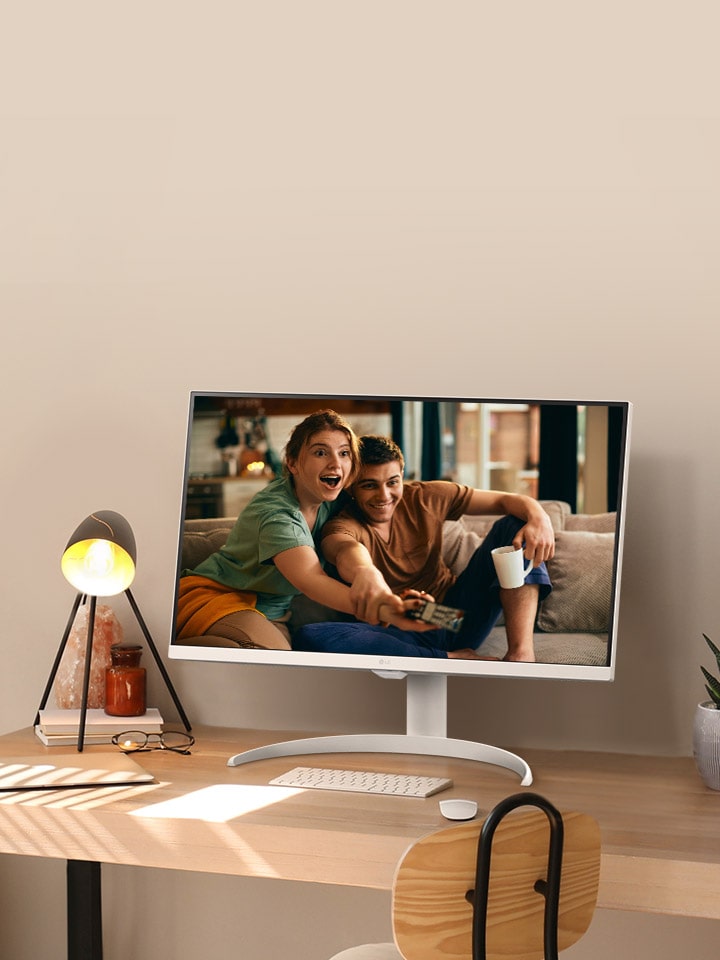 En el escritorio al lado de la ventana hay un LG MyView Smart Monitor que muestra una película en la pantalla. Al lado del monitor hay una lámpara y una maceta con una planta. En la mesa hay un teclado y un ratón.