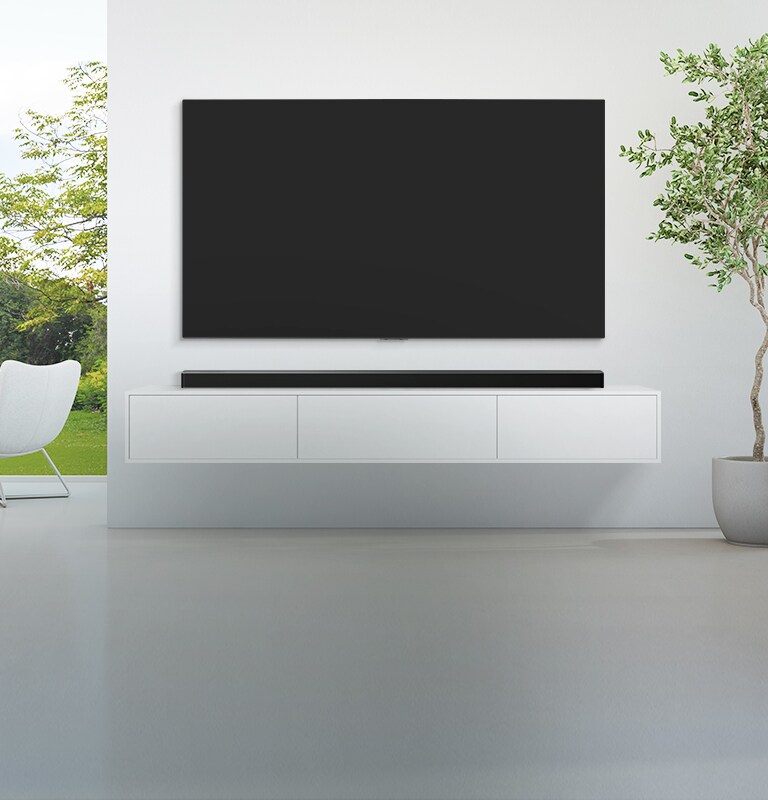 Un TV y una barra de sonido se han instalado en un salón amplio y blanco; desde la gran ventana se ve un bosque verde. 