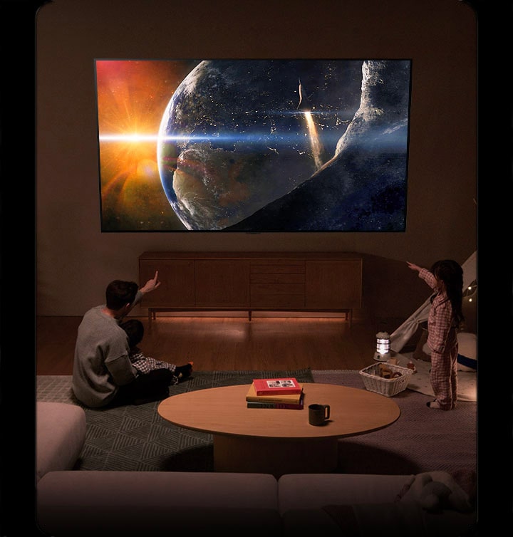 Une famille assise par terre dans une pièce à la lumière tamisée à côté d’une petite table, qui regarde un téléviseur LG installé sur le mur montrant la Terre depuis l’espace.