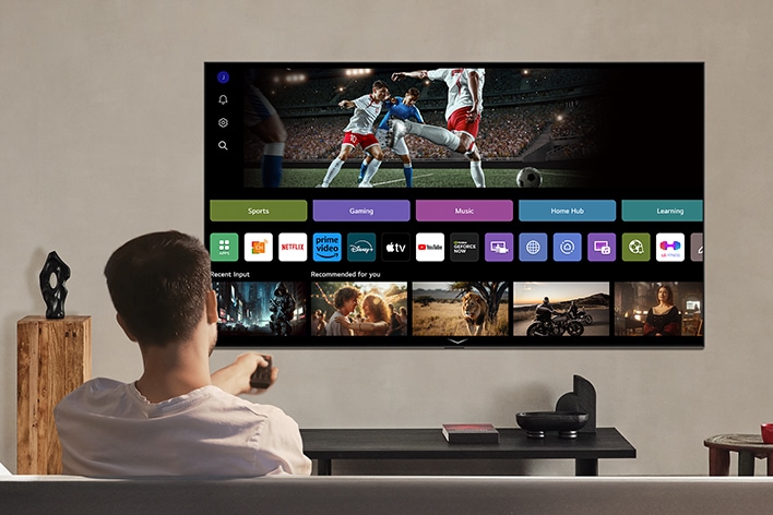 Une personne sur le canapé tenant une télécommande. LG AI TV sur le mur avec LG webOS à l’écran.