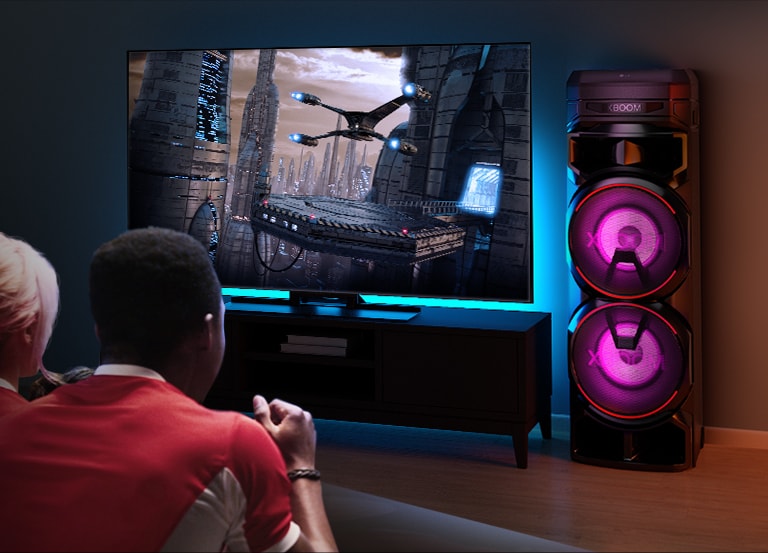 Des gens regardent la télévision sur un mur, avec une enceinte LG XBOOM RNC9 sur la droite. La télévision diffuse un film.