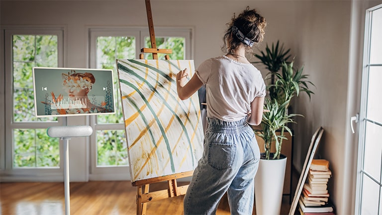 Le LG StanbyME est placé dans le salon avec le haut-parleur XT7S. Une femme dessine tout en écoutant de la musique sur l'écran et le haut-parleur.