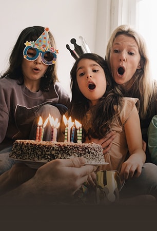 Image de deux femmes adultes et d’une jeune fille avec un chapeau d’anniversaire sur la tête et soufflant les bougies du gâteau.