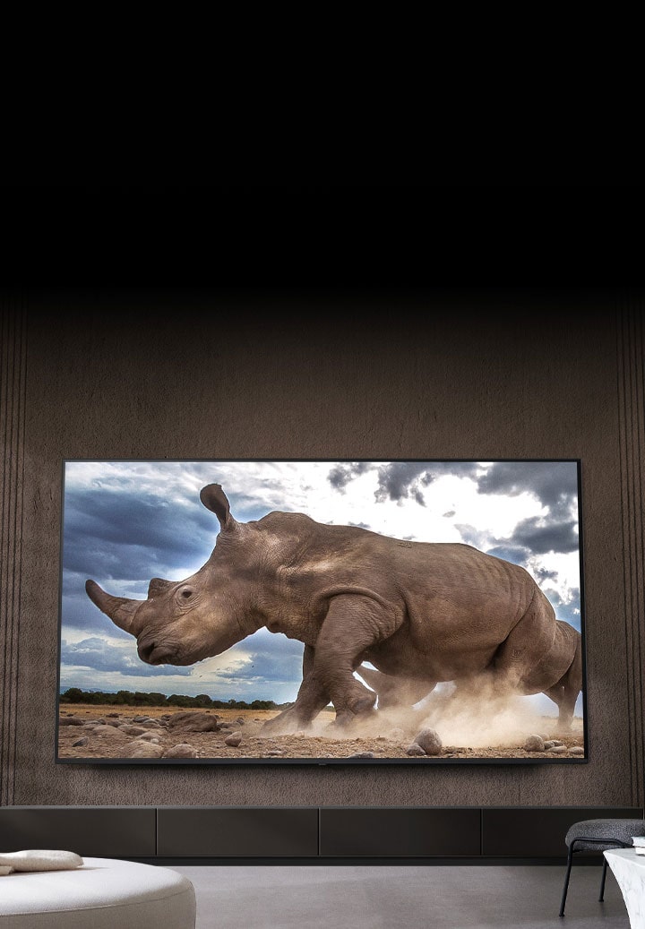 A rhinoceros in a safari setting is shown on an Ultra Big LG TV, mounted on the brown wall of a living room surrounded by cream-colored modular furniture.