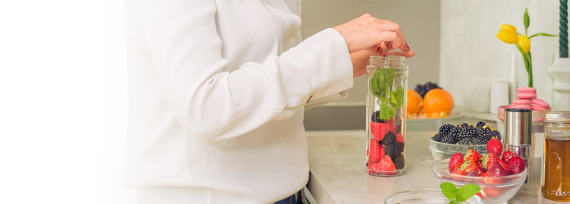 Une femme place des fraises, des myrtilles et des herbes dans un grand bocal en verre, entourée de bols en verre remplis d'ingrédients.