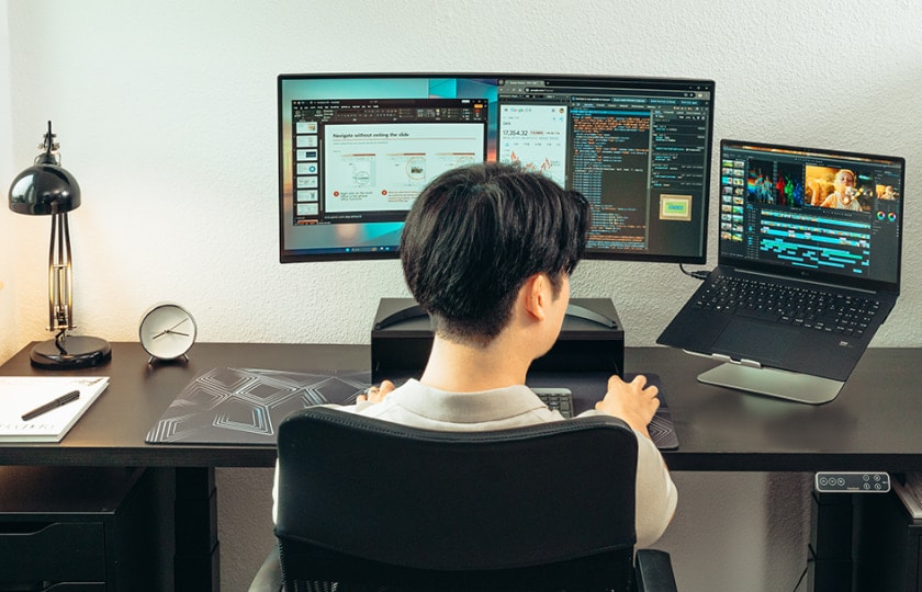 Vue de dos d'un homme à un bureau avec un large écran et un ordinateur portable, affichant des applications de codage et de productivité.