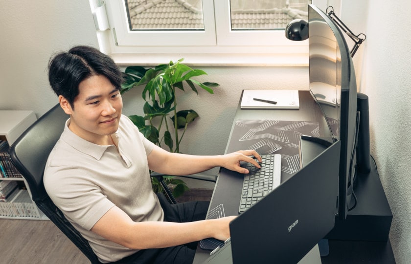 Homme assis à un bureau dans un bureau à domicile lumineux rempli de plantes, travaillant sur un ordinateur portable LG gram connecté à un moniteur externe.